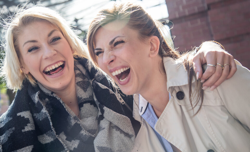 Zwei Frauen lachen herzlich und freuen sich gemeinsam in der Sonne, während sie sich arm in arm umarmen.