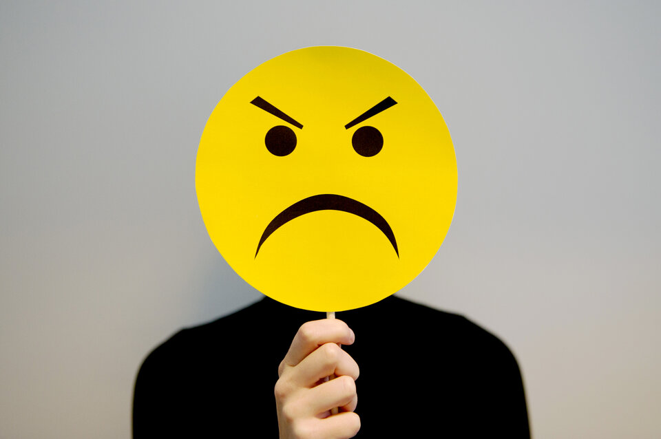 Eine Person hält ein gelbes Smiley-Schild mit wütendem Gesichtsausdruck vor ihr.