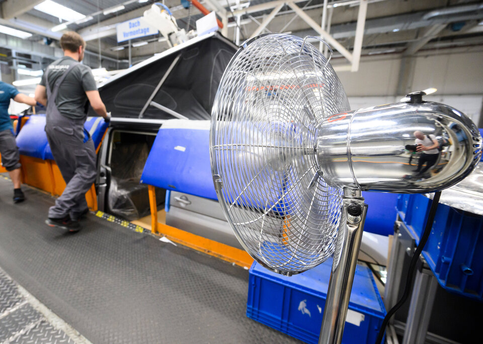 Ein industrieller Ventilator im Vordergrund, während Arbeiter an einer Maschine in einer Produktionshalle tätig sind.