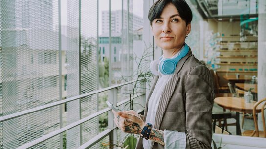 Eine Frau mit kurzen dunklen Haaren steht im Aufenthaltsbereich eines Büros. Sie hat am rechten Arm und Hand Tattoos und Armbänder um. Um den Hals hängen Kopfhörer.
