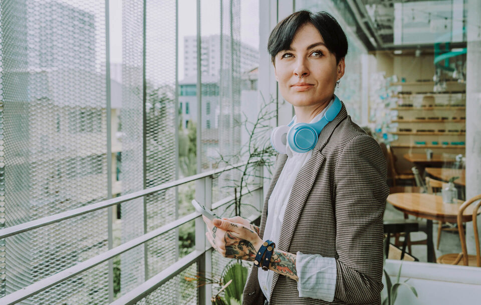Eine Frau mit kurzen dunklen Haaren steht im Aufenthaltsbereich eines Büros. Sie hat am rechten Arm und Hand Tattoos und Armbänder um. Um den Hals hängen Kopfhörer.