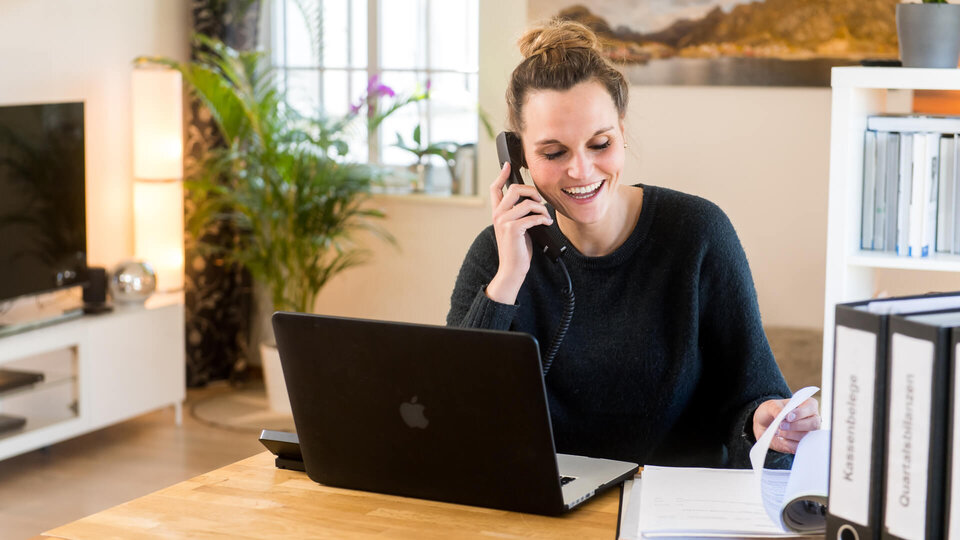 Eine Frau sitzt in ihrer Wohnung am Tisch mit einem Laptop und telefoniert. Beim Telefonieren lächelt sie.