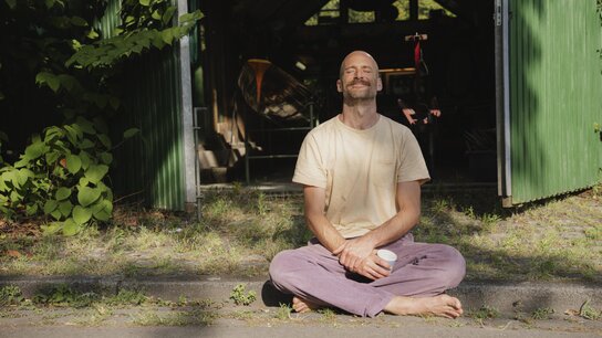 Ein Mann sitzt im Schneidersitz vor einer grünen Scheune, lächelt entspannt und hält eine Tasse in der Hand.