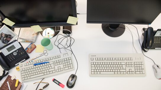 Zwei Computerarbeitsplätze mit zwei Monitoren, Tastaturen, Telefon, Bürobedarf und persönlicher Dekoration.