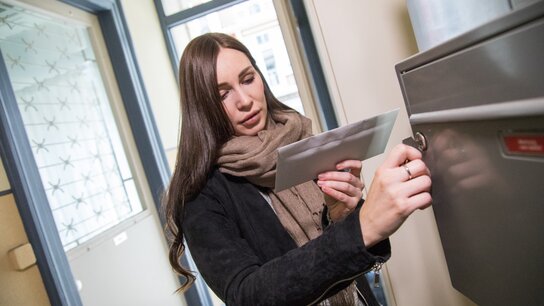 Eine Frau mit langen Haaren holt einen Brief in einen grauen Briefkasten in einem hellen Flur aus dem Briefkasten.