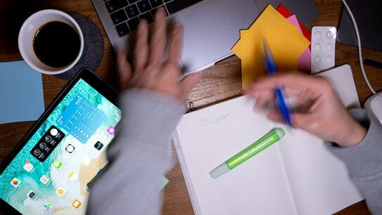 Ein Schreibtisch mit einem Laptop, einem Tablet, Notizbuch, bunten Zetteln und einer Tasse Kaffee. Hände schreiben und tippen.