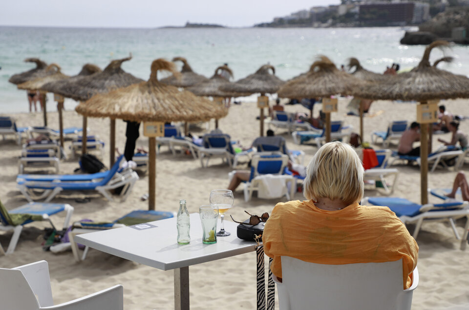 Eine Person sitzt im T-Shirt an einem Tisch mit Blick auf den Strand mit vielen Sonnenschirmen und dem Meer im Hintergrund.