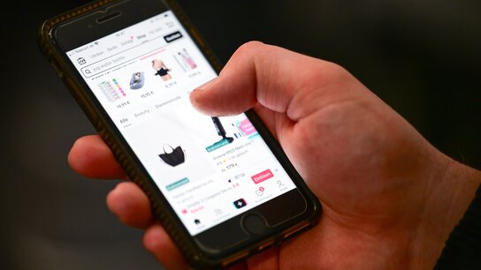 Ein Hand hält ein Smartphone, auf dem eine Shopping-App mit verschiedenen Produkten angezeigt wird.