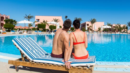 Paar sitzt auf einer Liege am Pool, im Hintergrund Palmen und sonnige Hotelanlage. Klare blaue Wasseroberfläche.