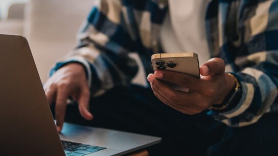 Person sitzt mit Laptop und Smartphone in der Hand, während er auf der Couch arbeitet.