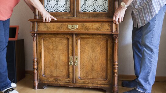 Zwei Personen heben gemeinsam ein detailliert verziertes Holzmöbel mit einer Glasfront und einem Oberteil mit Holzverzierungen.