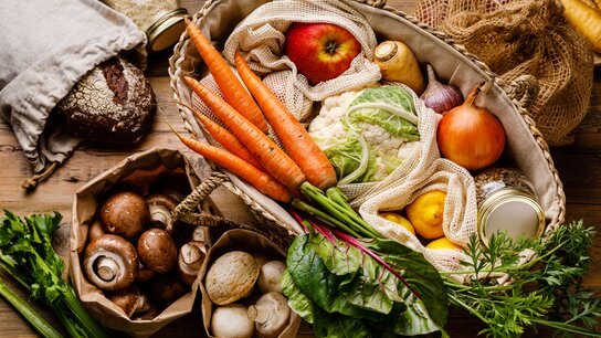 Ein Korb gefüllt mit frischen Gemüse, Äpfeln, Pilzen, Brot und weiteren Zutaten auf einer Holzoberfläche.