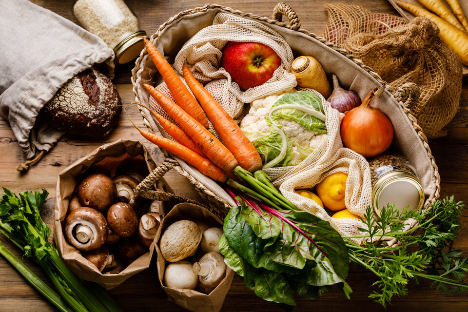 Ein Korb gefüllt mit frischen Gemüse, Äpfeln, Pilzen, Brot und weiteren Zutaten auf einer Holzoberfläche.