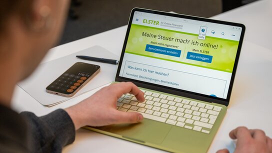 Arbeiter nutzt ein Laptop, um die ELSTER-Website für Online-Steuerverwaltung zu besuchen. Neben ihm liegt ein Taschenrechner.