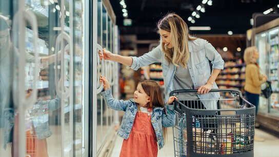 Eine Frau und ein kleines Mädchen stehen vor einem Kühlregal im Supermarkt. Das Mädchen zeigt auf ein Produkt.