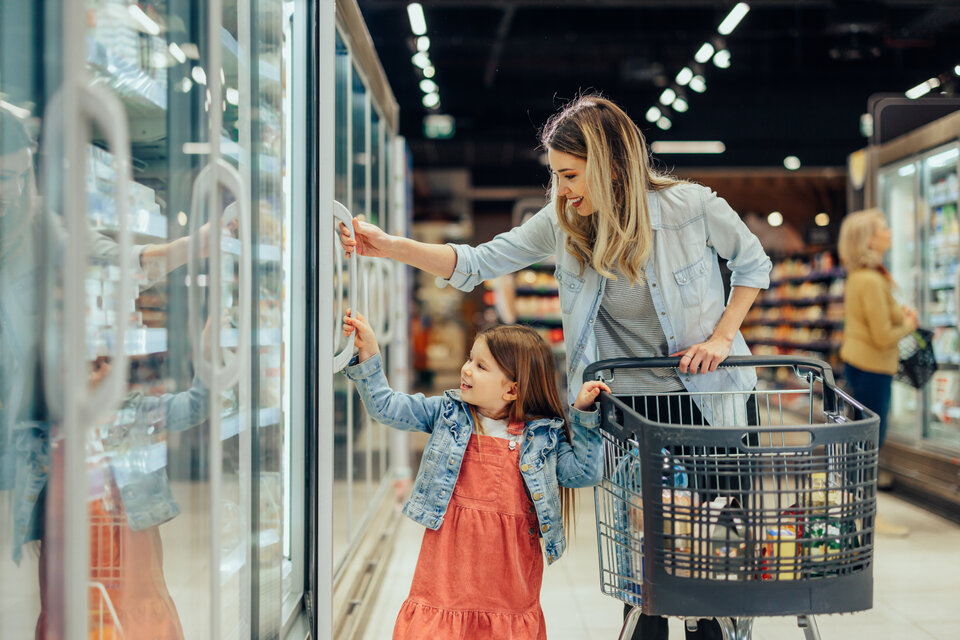Eine Frau und ein kleines Mädchen stehen vor einem Kühlregal im Supermarkt. Das Mädchen zeigt auf ein Produkt.