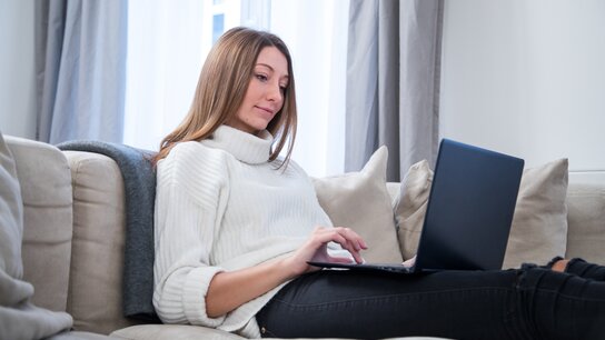 Eine Frau in einem hellen Pullover sitzt entspannt auf einem Sofa und arbeitet an einem Laptop.