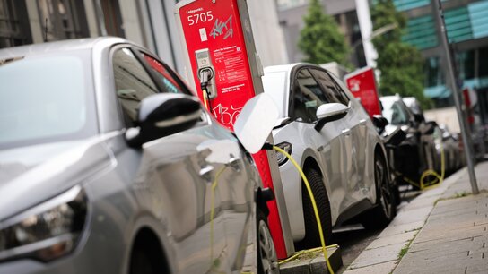 Zwei Elektroautos laden an einer roten Ladesäule, Kabel verbinden die Fahrzeuge mit der Station auf einer städtischen Straße.