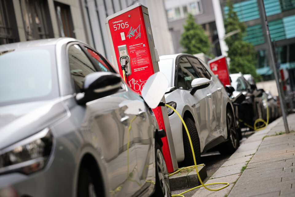 Zwei Elektroautos laden an einer roten Ladesäule, Kabel verbinden die Fahrzeuge mit der Station auf einer städtischen Straße.