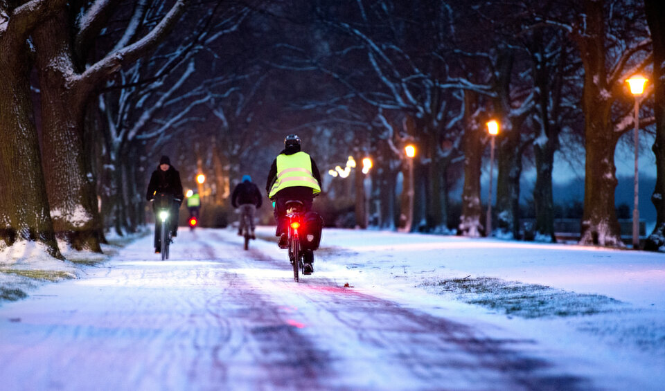 Radfahrer fahren in der Dämmerung durch eine schneebedeckte Allee, beleuchtet von Straßenlaternen.