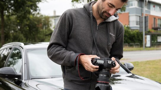 Mann hält eine Kamera in den Händen, steht neben einem schwarzen Auto, konzentriert auf das Display der Kamera.