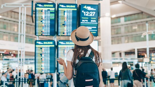 EIn Frau im Tshirt mit Hut auf steht in einer Abflughalle vor einer Infotafel.