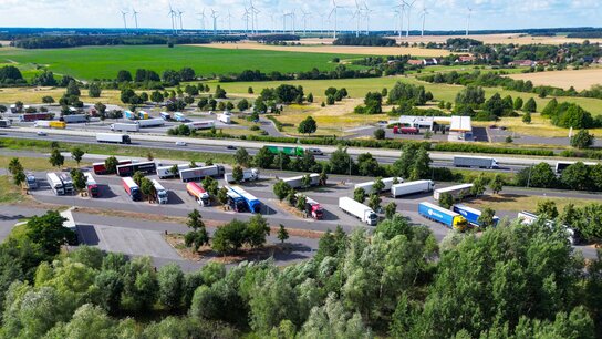 Luftaufnahme eines Parkplatzes mit zahlreichen Lkw, umgeben von Feldern, Bäumen und Windkraftanlagen im Hintergrund.