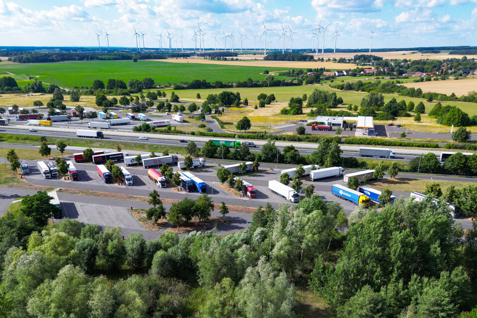 Luftaufnahme eines Parkplatzes mit zahlreichen Lkw, umgeben von Feldern, Bäumen und Windkraftanlagen im Hintergrund.