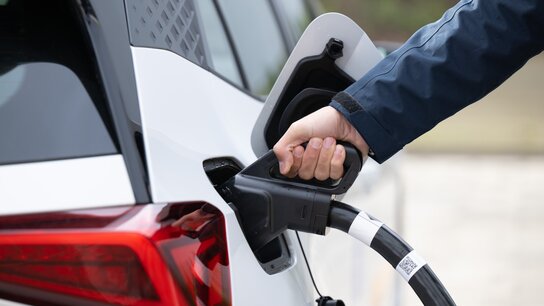 Eine Person hält einen Ladestecker, der in das Ladefeld eines weißen Elektroautos eingesteckt wird.