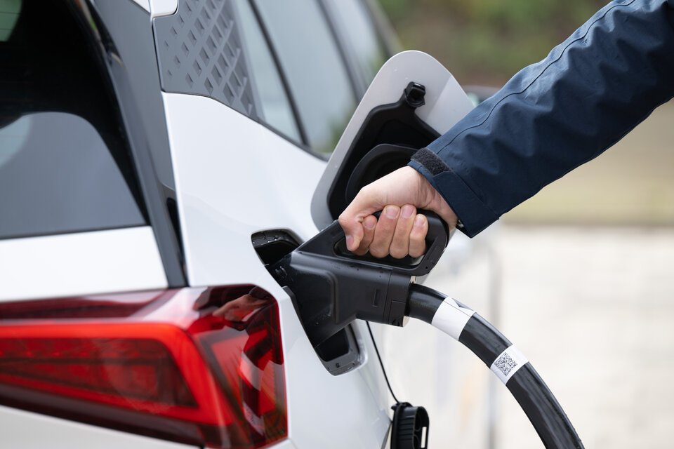 Eine Person hält einen Ladestecker, der in das Ladefeld eines weißen Elektroautos eingesteckt wird.