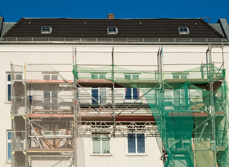 Fassade eines Gebäudes mit Baugerüst und grünen Netzabdeckungen, blauer Himmel im Hintergrund.