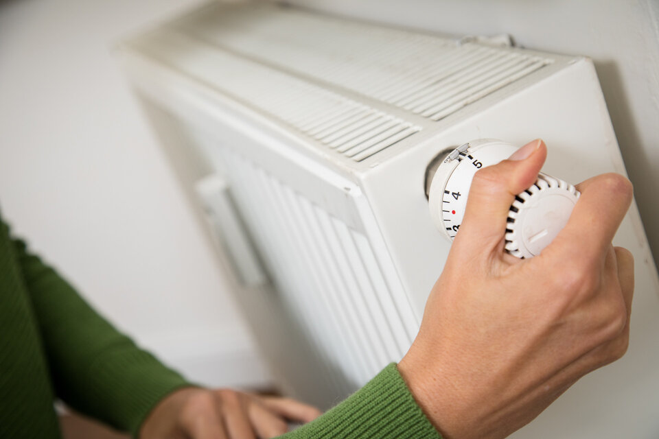 Hand dreht an der Thermostatregelung einer Heizkörperanlage in einem Wohnraum.