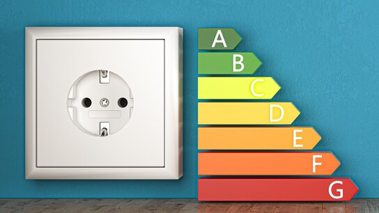 Steckdose an einer blauen Wand neben einer farbigen Energieeffizienzskala von A bis G auf Holzfußboden.