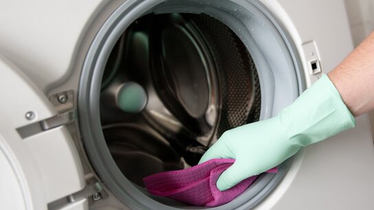 Hand mit grüner Gummihandschuh, die einen lila Reinigungstuch in die Öffnung einer Waschmaschine hält.