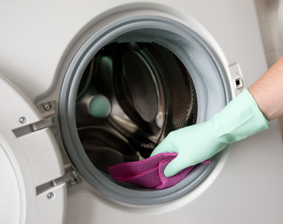 Hand mit grüner Gummihandschuh, die einen lila Reinigungstuch in die Öffnung einer Waschmaschine hält.