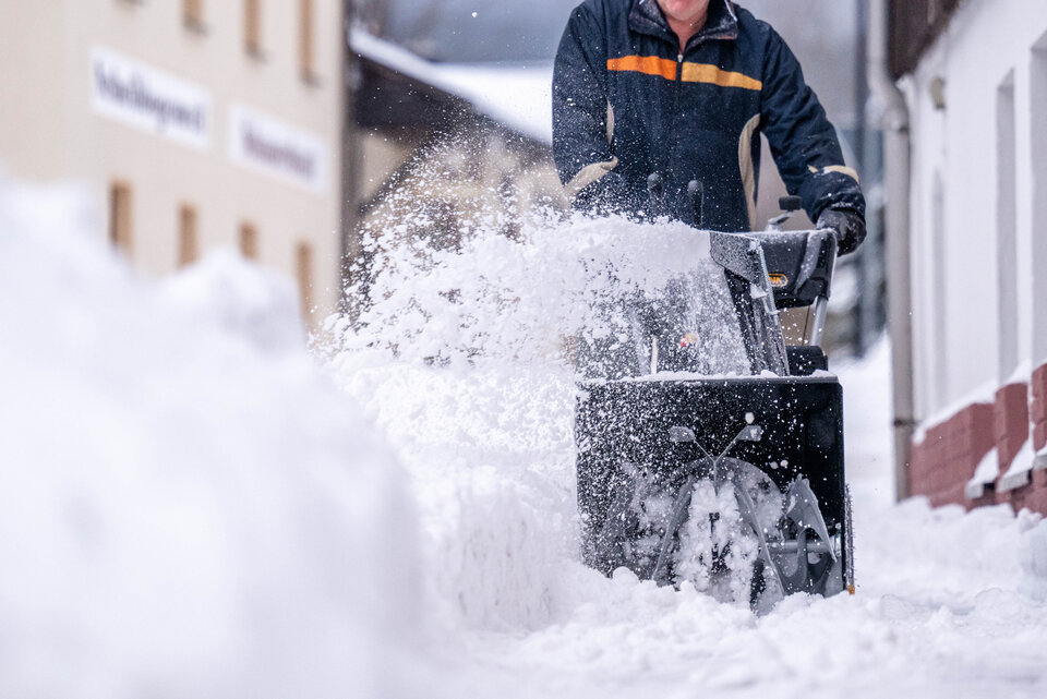 Mann räumt mit Schneefräse den schneebedeckten Bürgersteig frei, Schnee wird in die Luft geschleudert.