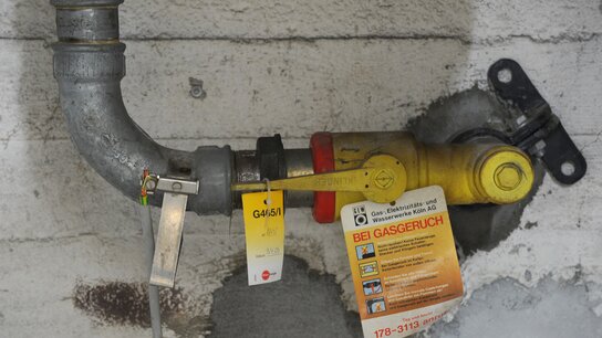 Gasventil mit einer gelben Kennzeichnung, an einer grauen Wand, mit Sicherheitsinformationen und einem Warnhinweis bei Gasgeruch.