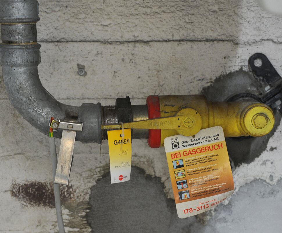 Gasventil mit einer gelben Kennzeichnung, an einer grauen Wand, mit Sicherheitsinformationen und einem Warnhinweis bei Gasgeruch.