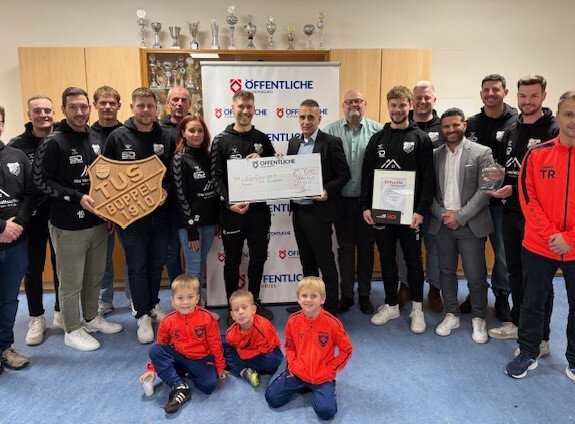 Gruppenfoto mit Sportteam und Unterstützern, die Pokale, Urkunden und ein Holzschild halten. Im Hintergrund Trophäen.