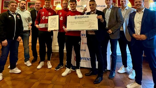 Gruppenszene mit 10 Personen, die einen Scheck und Urkunden für eine Auszeichnung präsentieren. Hintergrund in einem Veranstaltungsraum.