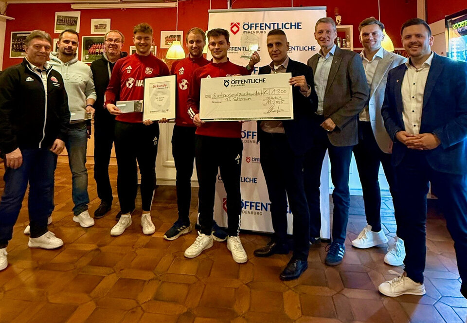 Gruppenszene mit 10 Personen, die einen Scheck und Urkunden für eine Auszeichnung präsentieren. Hintergrund in einem Veranstaltungsraum.