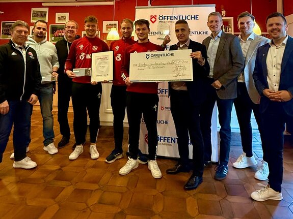 Gruppenszene mit 10 Personen, die einen Scheck und Urkunden für eine Auszeichnung präsentieren. Hintergrund in einem Veranstaltungsraum.