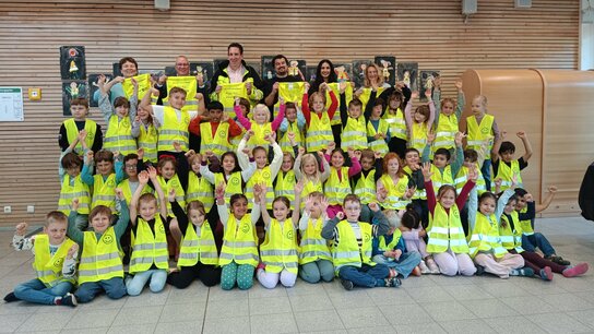 Gruppenfoto von Kindern in gelben Sicherheitswesten, die fröhlich die Arme heben, umgeben von Erwachsenen.