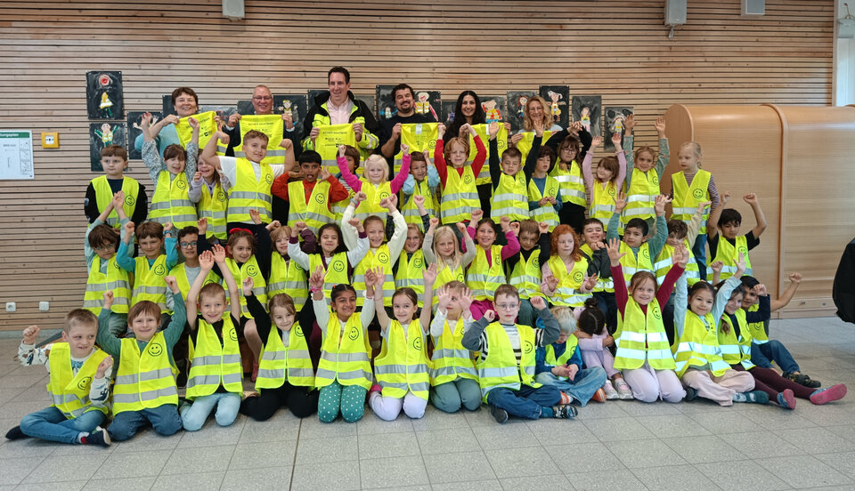 Gruppenfoto von Kindern in gelben Sicherheitswesten, die fröhlich die Arme heben, umgeben von Erwachsenen.