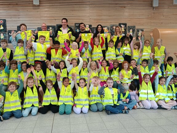 Gruppenfoto von Kindern in gelben Sicherheitswesten, die fröhlich die Arme heben, umgeben von Erwachsenen.