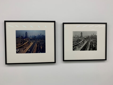 Zwei gerahmte Bilder an einer Wand: das linke in Farbe, das rechte in Schwarz-Weiß, beide zeigen eine Stadtansicht mit Verkehr.