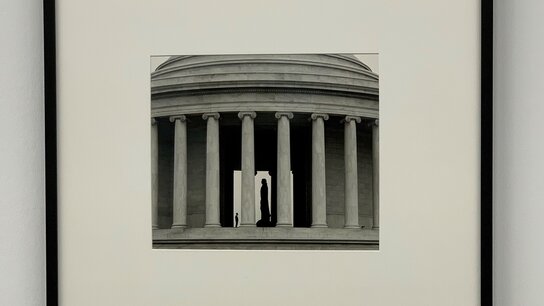 Silhouette einer Person vor architektonischen Säulen in Schwarz-Weiß, mit einer zentralen Öffnung im Gebäude.