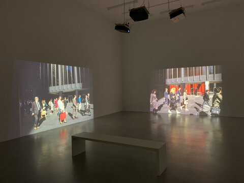 Zwei Projektionen in einem Ausstellungsraum zeigen Passanten in urbaner Umgebung, daneben eine weiße Bank.