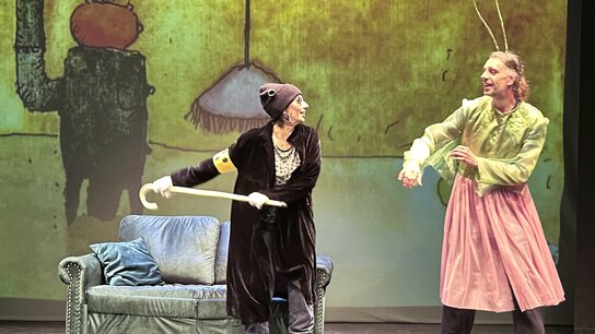 Zwei Personen in lustigen Kostümen stehen auf einer Bühne mit einem Sofa, während eine farbige Projektion im Hintergrund sichtbar ist.