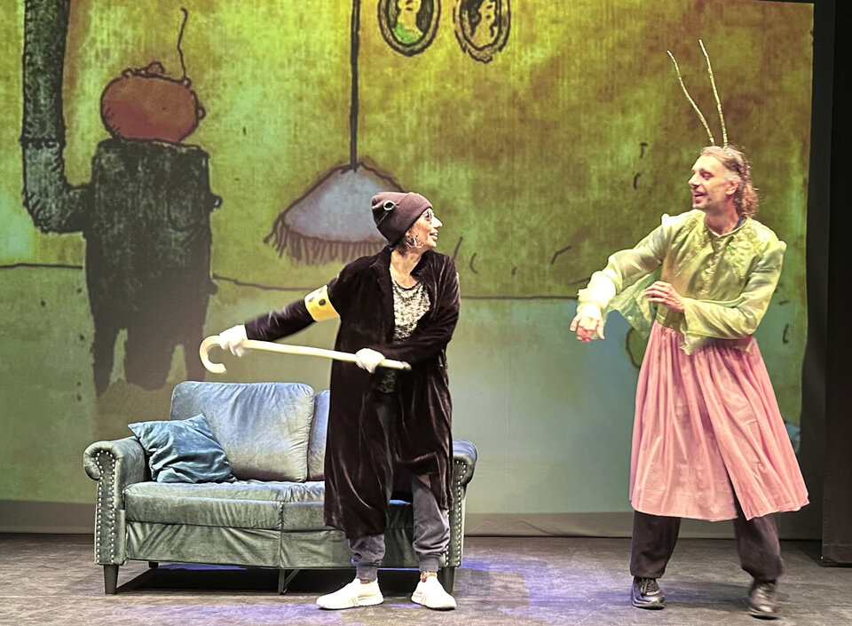 Zwei Personen in lustigen Kostümen stehen auf einer Bühne mit einem Sofa, während eine farbige Projektion im Hintergrund sichtbar ist.